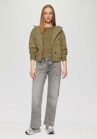 Olivgrüne Kapuzenjacke mit Fronttaschen, kombiniert mit einem taillierten olivgrünen Oberteil und grauen, weit geschnittenen Jeans. Silberne Sneakers vervollständigen das Outfit.