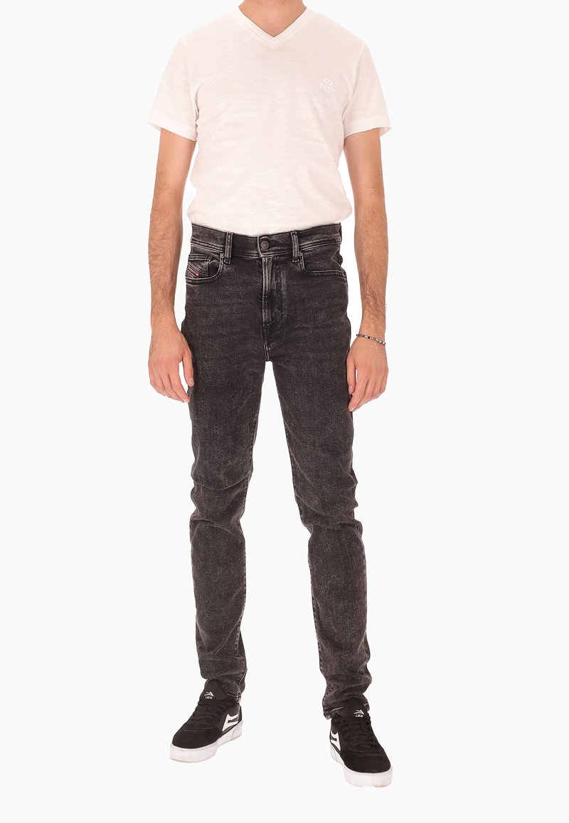 Homme portant un T-shirt à col en V blanc, un jean noir délavé et des baskets noires avec des semelles blanches, se tenant devant un fond blanc uni.