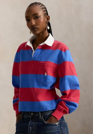 Κόκκινη και μπλε ριγέ μακρυμάνικη πόλο με λευκό γιακά και τσέπη στο στήθος. Διακοσμημένη με μικρό έμβλημα στην τσέπη. Φορεμένη με τζιν παντελόνι.