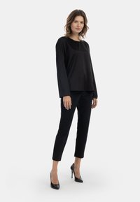 Donna con capelli medi, che indossa una maglietta nera a maniche lunghe, pantaloni neri attillati e tacchi alti neri, in piedi su uno sfondo chiaro e uniforme.