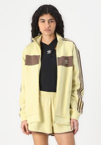 Yellow track jacket with brown stripes, zip front, and fitted cuffs, paired with a black top and matching shorts. Smooth fabric with logo accents.