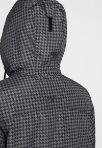 Veste à capuche avec motif pied-de-poule gris et noir, ornée d'un petit logo noir brodé sur le haut du dos.