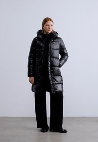 Μαύρο μπουφάν με καπιτονέ οριζόντια ραφή, καπότα, γυαλιστερό φινίρισμα και μήκος μέχρι το γόνατο. Συνδυασμένο με μαύρο σύνολο και παπούτσια.