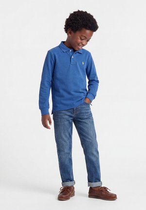 Jongen draagt een blauw poloshirt met lange mouwen, opgerolde blauwe spijkerbroek en bruine schoenen, glimlacht en kijkt naar beneden met één hand in zijn zak.