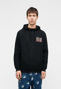 Mustaharma huppari, jossa on etutasku ja nyöripää, jossa vasemmassa rinnassa on värikäs graafinen painatus. Yhdistetty sinisiin farkkuihin, joissa on yin-yang-kuvioita.