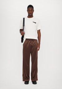 Weißes T-Shirt mit schwarzem Aufdruck, braune Satin-Hosen und schwarze Schuhe. Das Model hält eine schwarze Schultertasche. Einfaches, modernes Design.