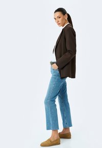 Blazer marrón sobre una blusa blanca, combinado con jeans azul claro y mocasines color tabaco. El conjunto presenta un diseño ajustado y jeans de pierna recta.