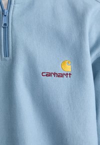 Sudadera de forro polar azul claro con media cremallera. Logo de Carhartt bordado en amarillo y rojo en el pecho. Textura suave, diseño casual.