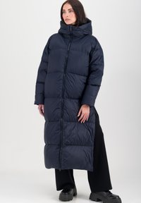 Langs blauwe pufferjas met een capuchon, voorzien van een ritssluiting en een gelaagd vullingdesign. Draagt over zwarte wijde broekspijpen en chunky laarzen.