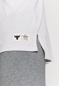 Bílé tričko s krátkým lemem, na kterém je štítek s grafikou býka a textem. Kombinováno se šedými legínami, které zdůrazňují texturu látky.