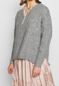 Grauer Strickpullover mit V-Ausschnitt, ausgestattet mit einem Zopfmuster und einem lockeren Schnitt. Wird über einem pinken, bedruckten Kleid mit rüschiniertem Saum getragen.