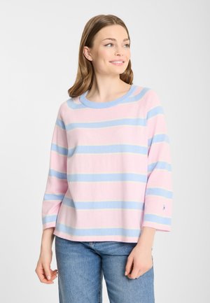 Jonge vrouw die lacht, draagt een roze en lichtblauw gestreepte trui en een blauwe spijkerbroek, kijkt iets naar rechts tegen een effen achtergrond.