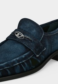 Mocassin bleu foncé à texture denim avec un emblème logo métallique argenté sur la bride et une semelle en caoutchouc noire.