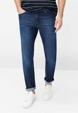 Mann trägt dunkelblaue hochgekrempelte Jeans, weiße Sneakers und ein schwarz-weiß gestreiftes Hemd, steht mit einer Hand in der Hosentasche.