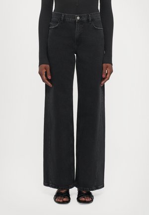 Persoon draagt zwarte wijde jeans, een zwart top met lange mouwen en zwarte sandalen met open tenen en kruisbanden.