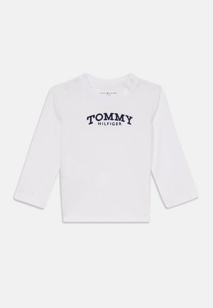 Camiseta blanca de manga larga para niño pequeño con texto en azul marino "Tommy Hilfiger" en el pecho y botones a presión en el hombro.