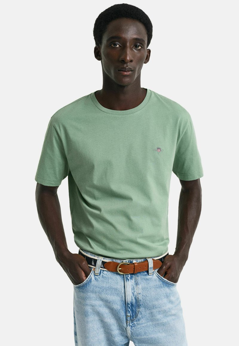 Hellgrünes Baumwoll-T-Shirt mit rundem Halsausschnitt und kurzen Ärmeln, verziert mit einem kleinen gestickten Logo auf der Brust. Dazu kombiniert mit hellblauen Jeans.