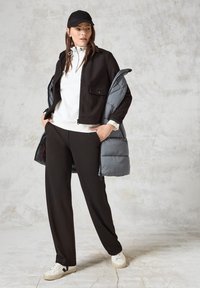 Giacca nera con zip e tasca frontale, felpa bianca con coulisse, pantaloni neri, gilet grigio imbottito appeso e sneakers bianche.