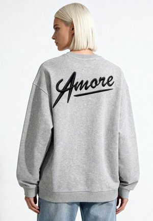 Person mit kurzen blonden Haaren, die einen grauen Pullover mit schwarzem "Amore" Schriftzug auf dem Rücken trägt, kombiniert mit hellblauen Jeans.