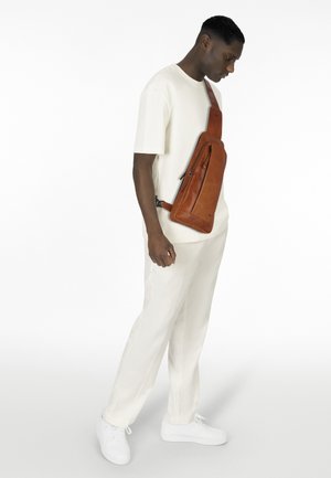 Borsa a tracolla in pelle marrone dal design elegante, dotata di tasca con zip e tracolla regolabile, indossata da una persona in un outfit bianco e sneakers.