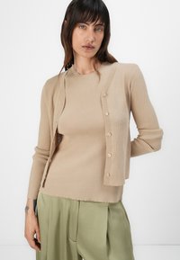 Beige gerippter Cardigan mit rundem Halsausschnitt, Knopfleiste und langen Ärmeln, über einem passenden beige Oberteil getragen. Weiches Strickmaterial.
