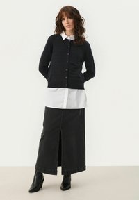 Cardigan negru cu nasturi peste o cămașă albă, asociat cu o fustă lungă din denim negru, cu o deschidere frontală. Botine negre, la gleznă, completează ținuta.