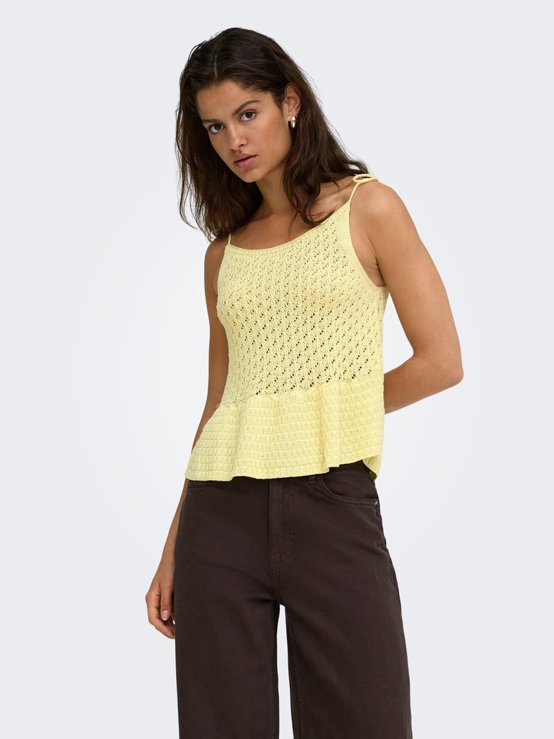 Femme portant un haut en maille jaune clair sans manches avec un bas à basque et un pantalon marron foncé taille haute sur un fond clair uni.