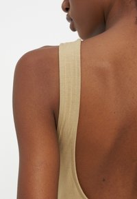 Κοντινή λήψη του ώμου και του άνω μέρους της πλάτης ενός ατόμου που φοράει μπεζ αμάνικο τοπ με χοντρό λουράκι, σε απλό ανοιχτό φόντο.