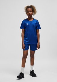 Blå sportuniform av slät, lätt tyg. Inkluderar en V-ringad tröja och shorts med svarta axelaccenter. Svarta sneakers och vita strumpor.