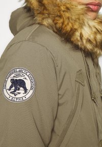 Parka verde oliva con cappuccio foderato in pelliccia, dotata di una toppa circolare con un design di orso sulla manica e di un cordoncino sul davanti.