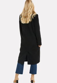 Cappotto lungo nero con taglio dritto e spacco posteriore, abbinato a jeans blu con orli sfrangiati e stivaletti con tacco marrone chiaro.
