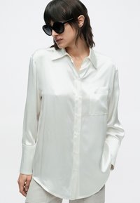 Persoană cu păr închis la culoare, purtând ochelari de soare negri oversized și o cămașă albă din satin cu mâneci lungi, nasturi și buzunar frontal.