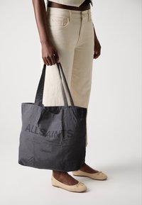 Melna auduma soma ar garām rokturiem, uz kuras izdrukāti vārdi "ALLSAINTS" un "LONDON" treknā fontā. Gluda tekstūra, vienkāršs dizains.