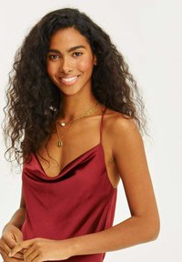 Bordová saténová blúzka s drapériou na výstrihu, tenkými špagetovými ramienkami a hladkou textúrou. Obsahuje vrstvené zlaté náhrdelníky s perlami.