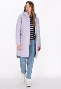 Lichtpaarse lange jas met kap, gemaakt van een gladde stof. Gecombineerd met een zwart-wit gestreepte top en blauwe spijkerbroek. Witte sneakers zichtbaar.
