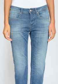 Cintura y piernas de una persona que lleva jeans azules descoloridos de subida media con bolsillos frontales y cierre de botón, manos relajadas a los lados.