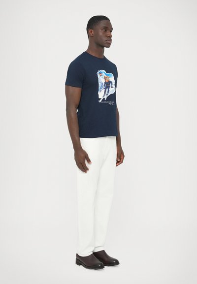 Teget pamučna majica s grafičkim dizajnom sprijeda, uparena s bijelim hlačama i tamnim cipelama. Model stoji u neutralnom položaju.