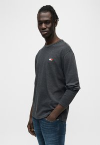 Γκρι μακρυμάνικη μπλούζα με στρογγυλή λαιμόκοψη, με μικρό πολύχρωμο λογότυπο στο στήθος. Φορεμένη με στενά μπλε τζιν.