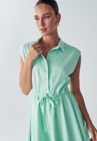 Mint zelena brezrokavna obleka iz mehkega blaga, ki ima zadrgo spredaj, ovratnik in regulirajoč pas z vrvico.