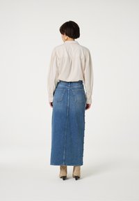 Jeans-Maxi-Rock mit geradem Schnitt in hellblauer Farbe, ausgestattet mit Gesäßtaschen und seitlichen Knopfdetails zum Verschließen. Kombiniert mit einer beigen Bluse.
