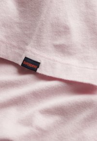 Světle růžové tričko vyrobené z měkké látky, s malou tmavě modrou cedulkou s nápisem "SUPERDRY" v červeném písmu. Hladká textura, design s krátkým rukávem.