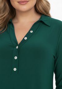 Chemise à boutons vert foncé avec un col et cinq boutons blancs le long de la patte de boutonnage. Tissu lisse avec un léger éclat et un design ajusté.