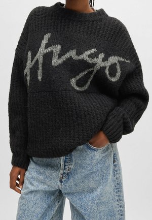 Personne portant un pull en tricot texturé noir avec un grand texte manuscrit blanc "Kenzo" associé à un jean en denim bleu clair, main dans la poche.