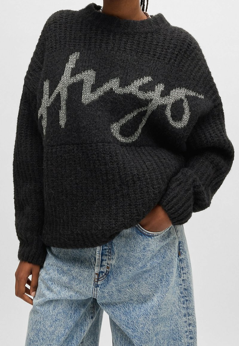 Personne portant un pull en tricot texturé noir avec un grand texte manuscrit blanc "Kenzo" associé à un jean en denim bleu clair, main dans la poche.