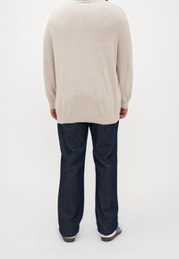 Pull beige en tricot à manches longues, bordure noire autour du col, associé à un pantalon en denim bleu foncé et des chaussures noires à rayures blanches.