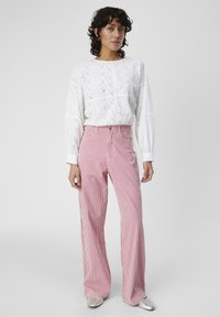 Blouse blanche à manches longues avec un motif à œillets, associée à un pantalon large à rayures rouges et blanches, complété par des chaussures argentées brillantes.
