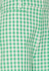 Zelená a bílá látka se vzorem kostiček, tvořená mřížkovým designem s malými čtverci. Textura působí tkaně. Viditelný je detail ploché kapsy.