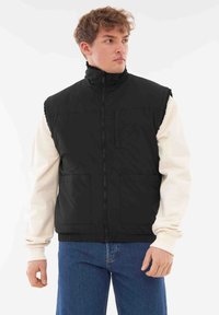 Fekete pufferszatyor magas gallérral, cipzárral és két elülső zsebbel, egy krémszínű pulóver felett viselve, kék farmernadrággal párosítva.