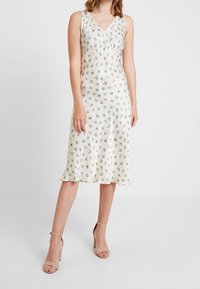 Robe A-line sans manches en tissu crème clair, présentant un motif floral en vert et bleu, avec un col en V et un ourlet festonné.