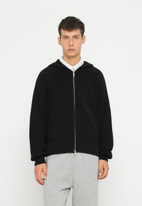 Svart hettegenser med glidelås, med ribbet tekstur, løs passform og vide ermer. Kombinert med grå joggebukser.
